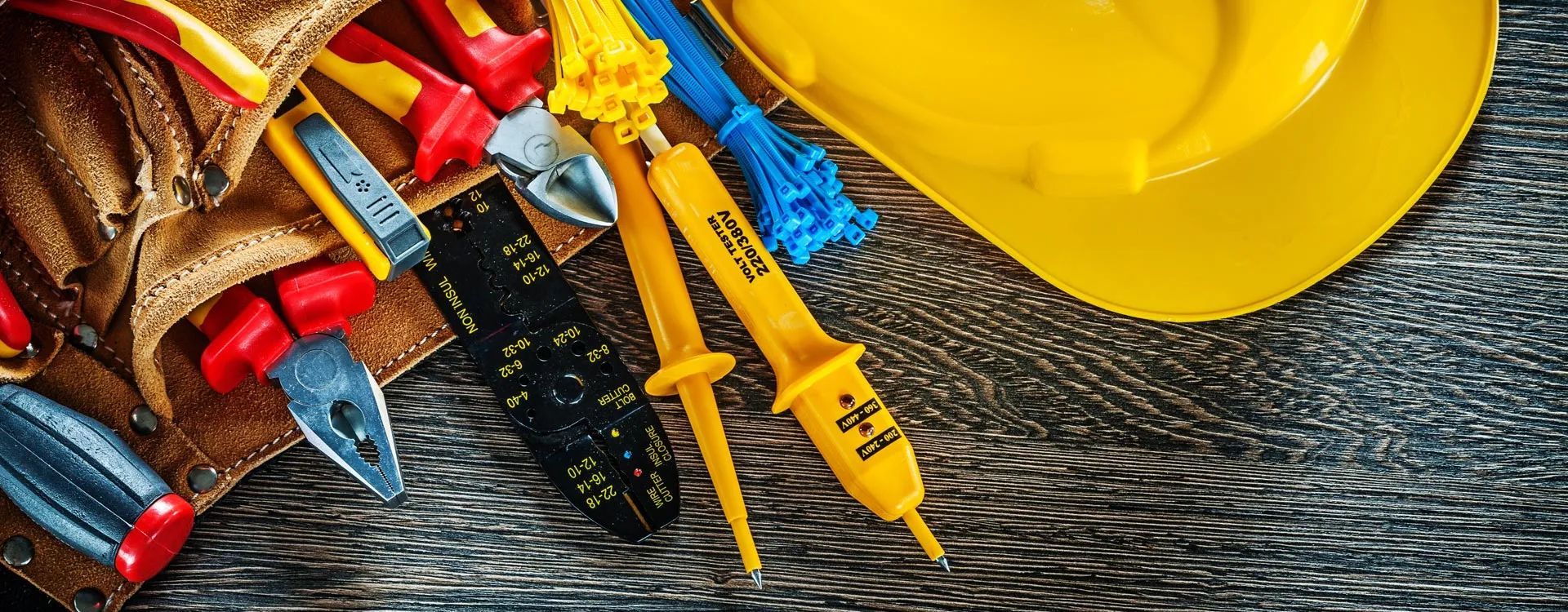 Tools of an electrician on a wooden surface, including a hard hat and a tool belt with pliers and a voltage tester.