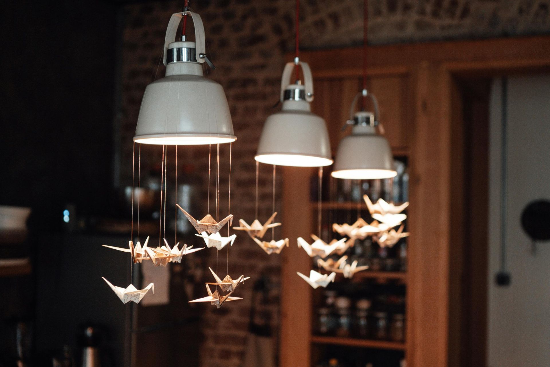 Three industrial pendant lights hang over paper origami cranes suspended by thin strings in a rustic kitchen.