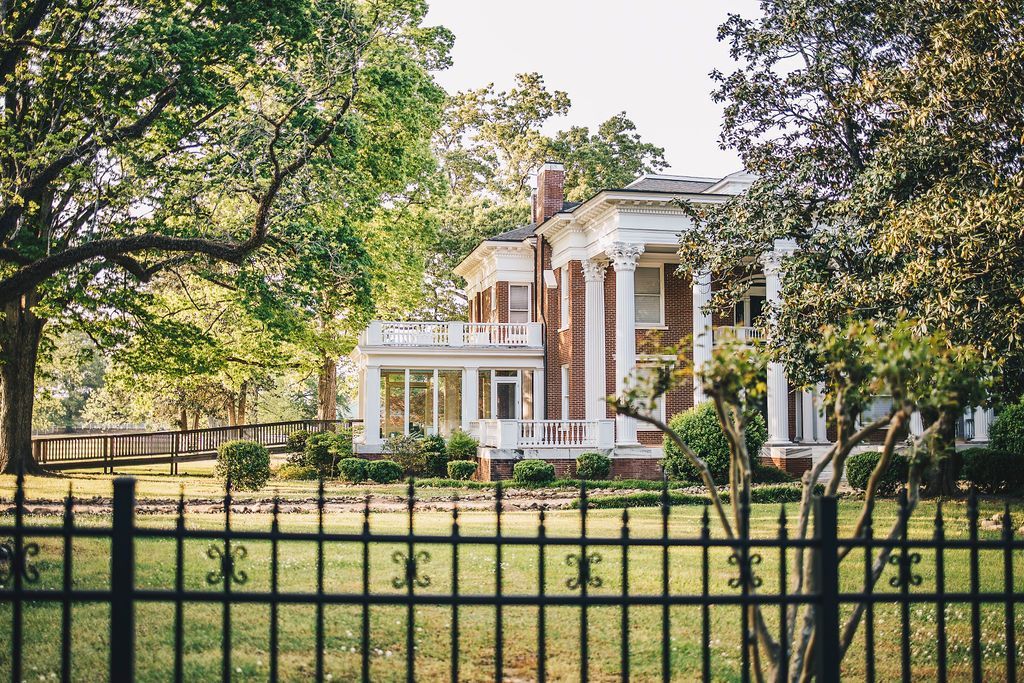 A large white house is behind a black fence surrounded by trees.