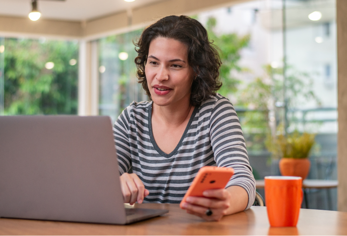 Uma mulher está sentada à mesa usando um laptop e um celular.