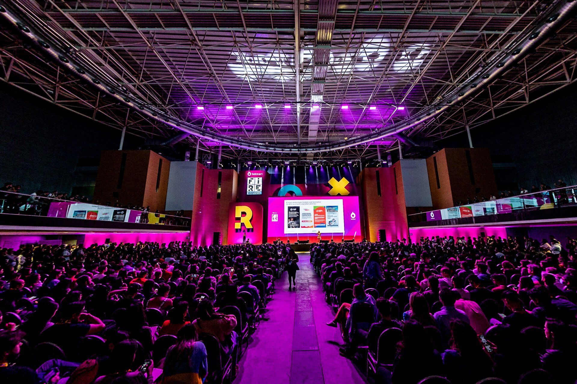 Uma grande multidão de pessoas está sentada em frente a um palco com luzes roxas.