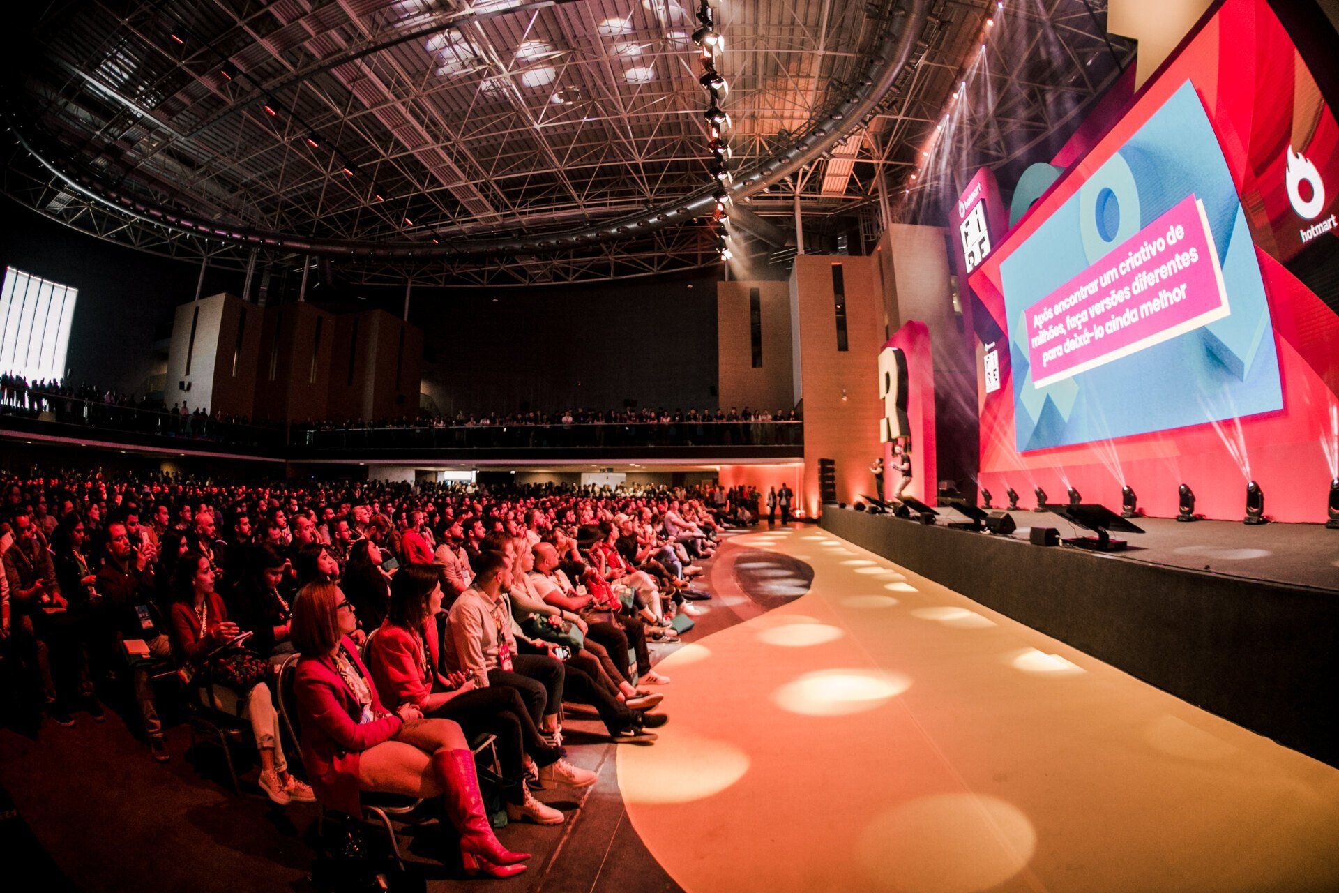 Uma grande multidão de pessoas está sentada em um grande auditório assistindo a uma apresentação.