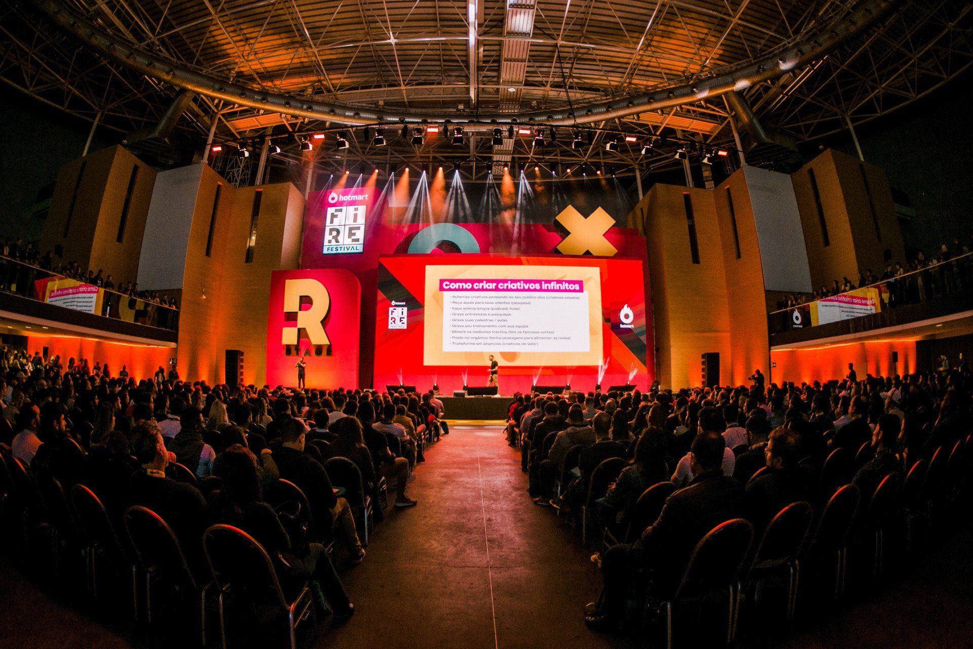 Uma grande multidão de pessoas está sentada em um grande auditório assistindo a uma apresentação.