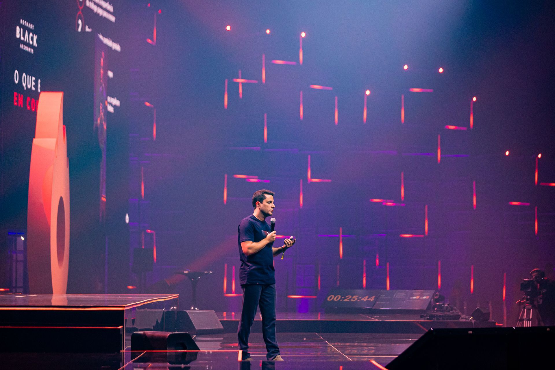 Foto do CEO e confundador da Hotmart, João Pedro Resende, palestrando no palco I do Hotmart FIRE 2024. Ele está de roupas pretas, com um microfone na mão, e o palco tem tons de roxo e rosa na iluminação