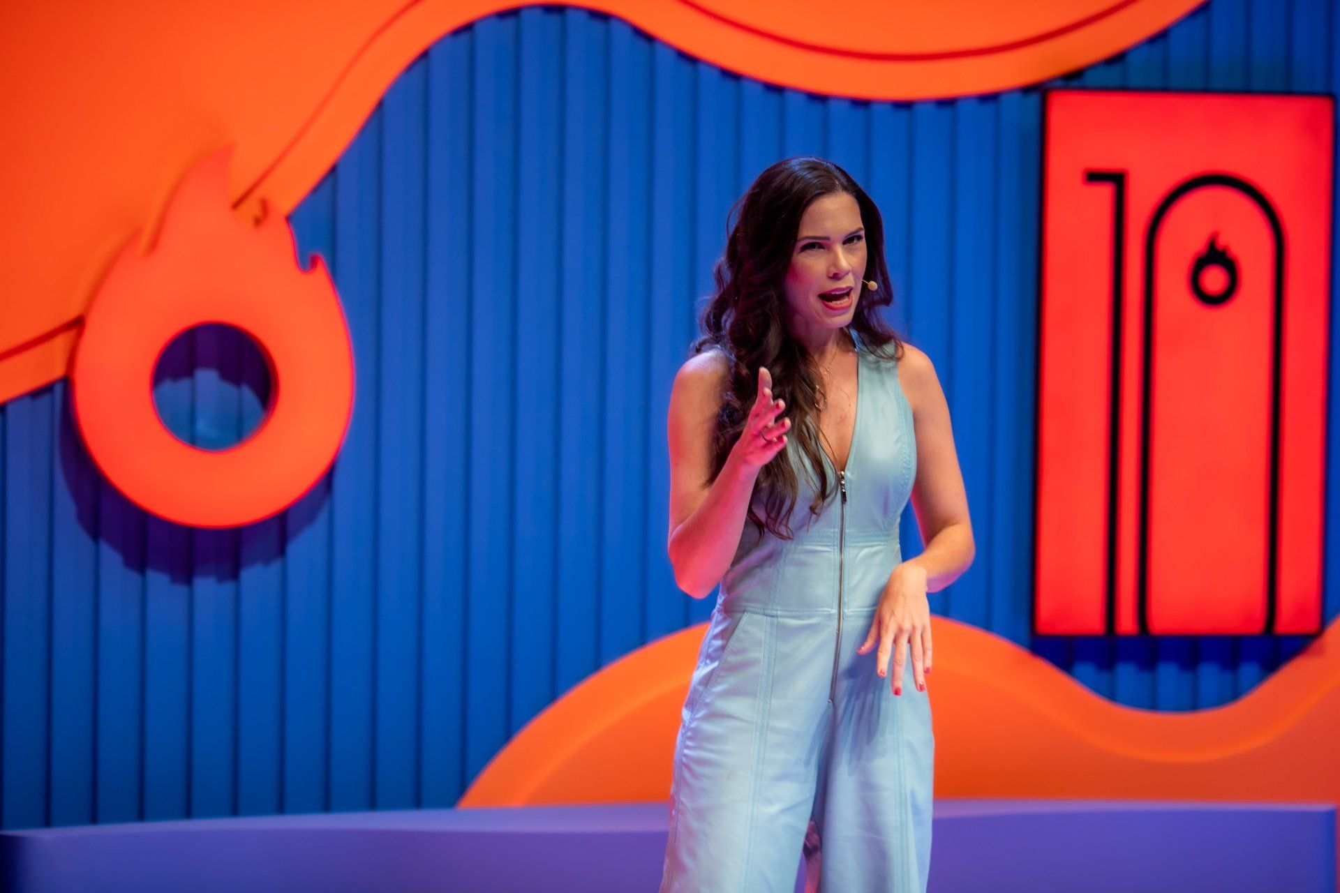 Uma mulher de macacão azul está parada em frente a um microfone no palco.