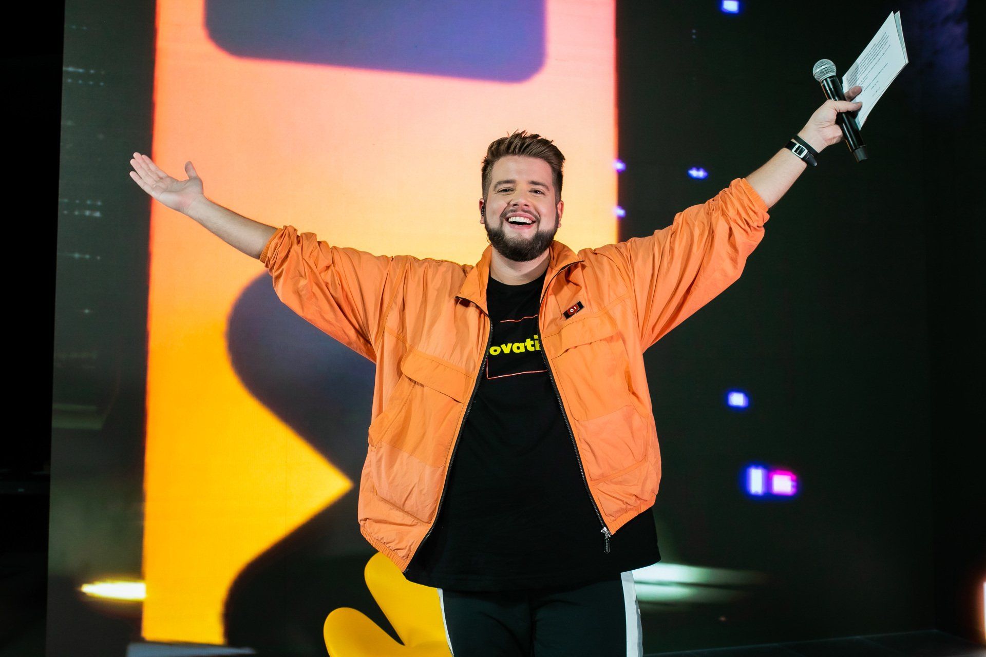 Um homem de jaqueta laranja segura um microfone em frente a uma tela grande.