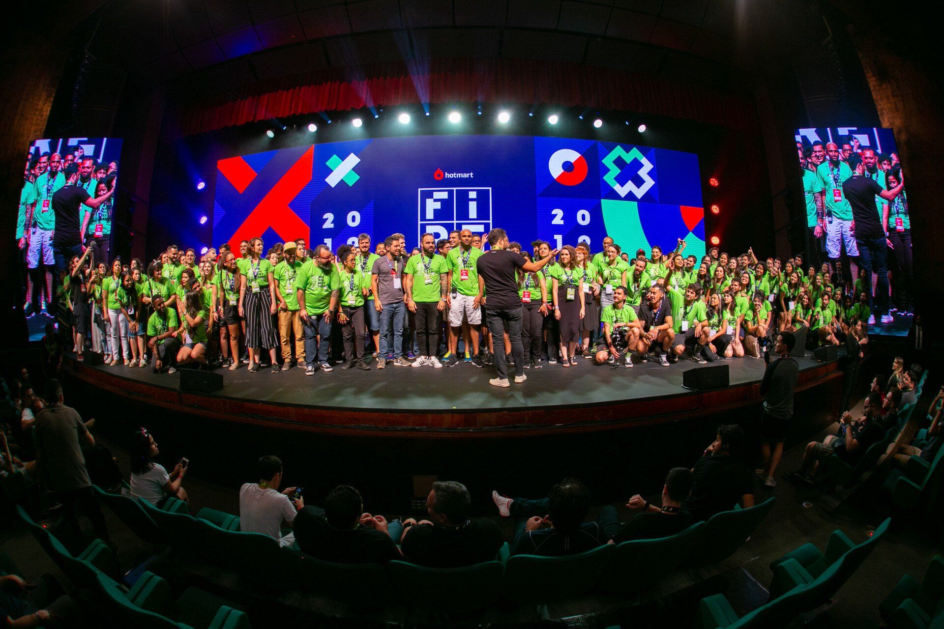 Um grande grupo de pessoas está em pé em um palco em frente a uma tela grande.