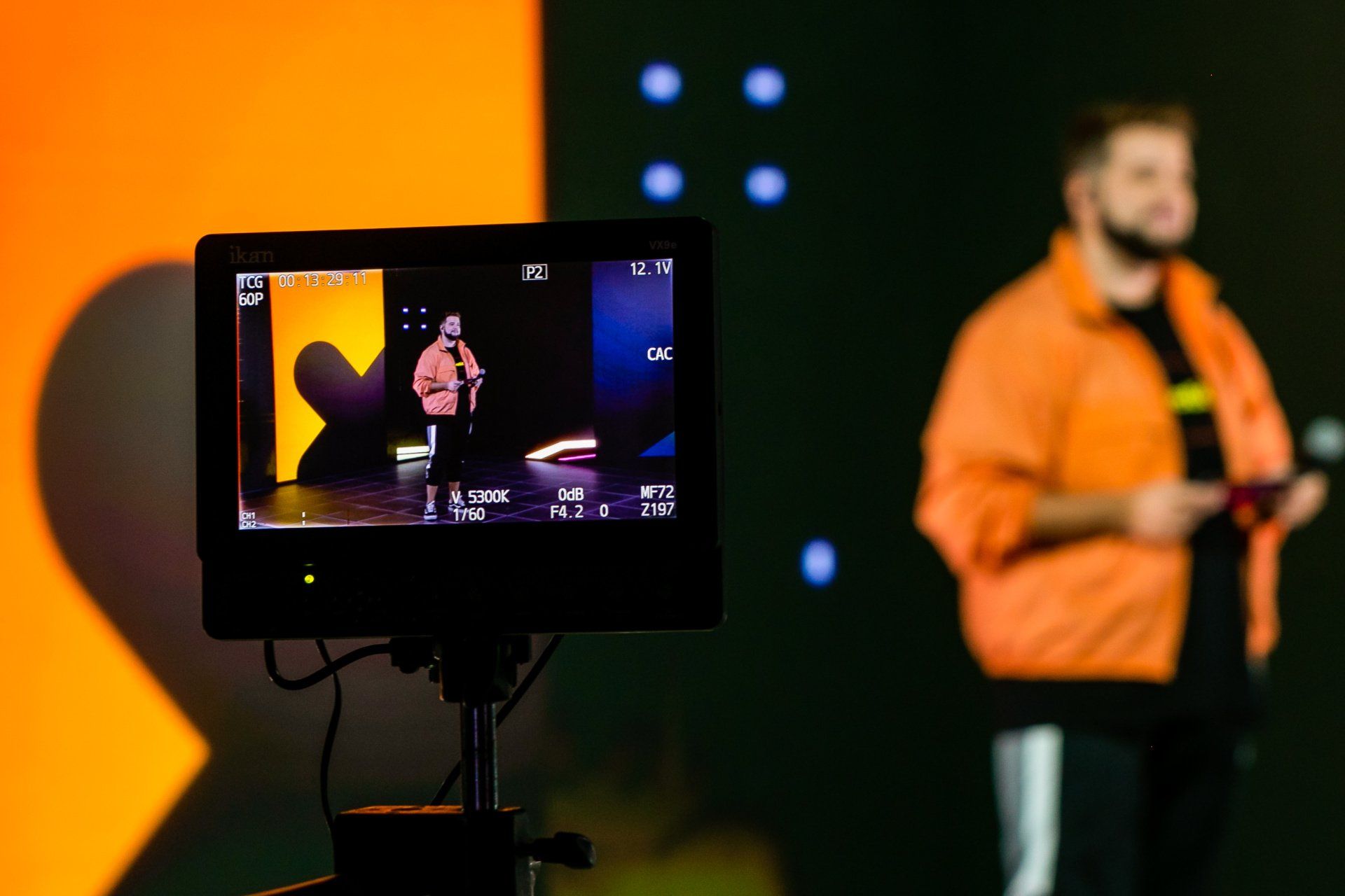 Um homem com uma jaqueta laranja está parado na frente de uma câmera.