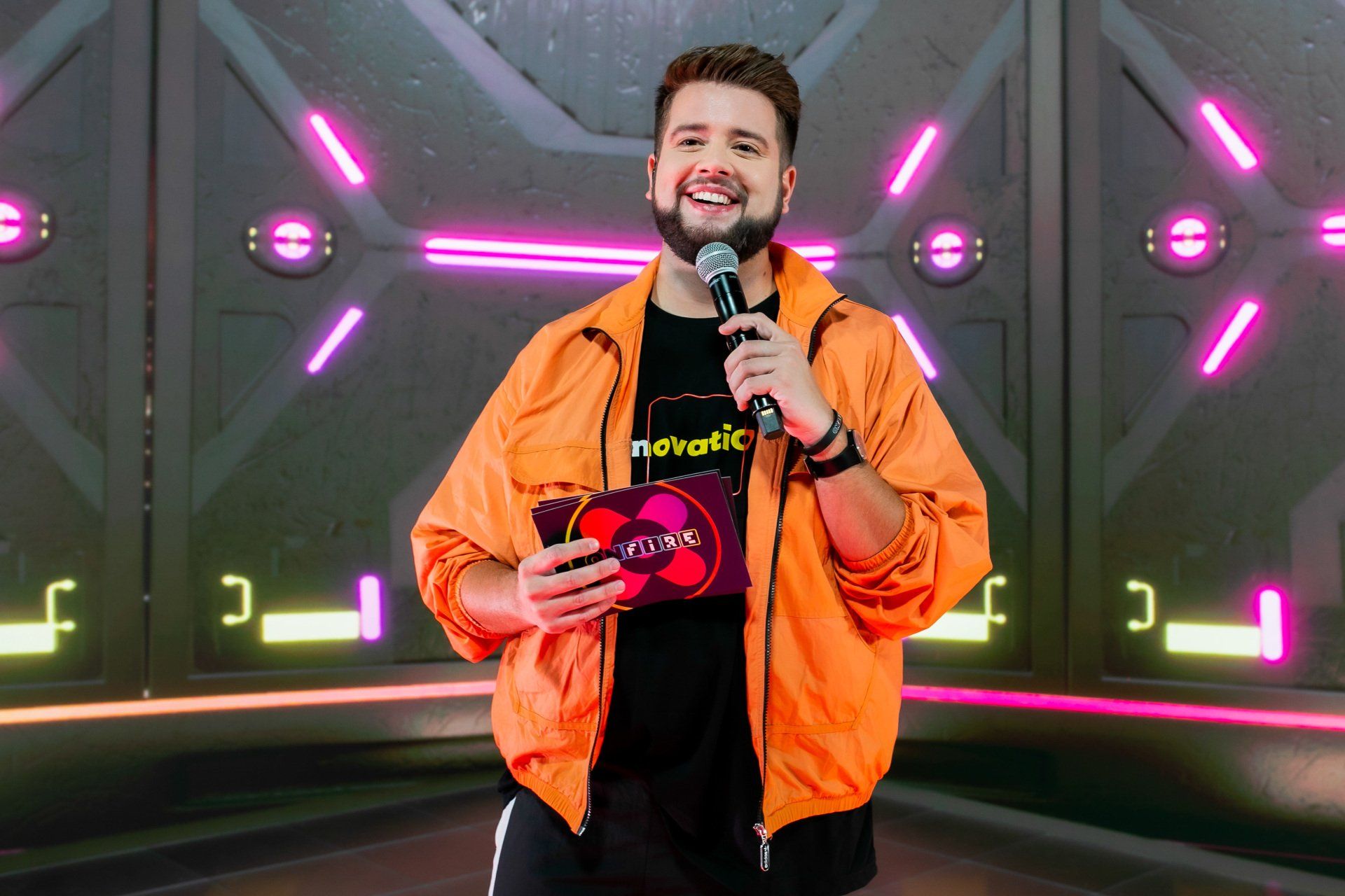 Um homem de jaqueta laranja está segurando um microfone em frente a uma parede de neon.