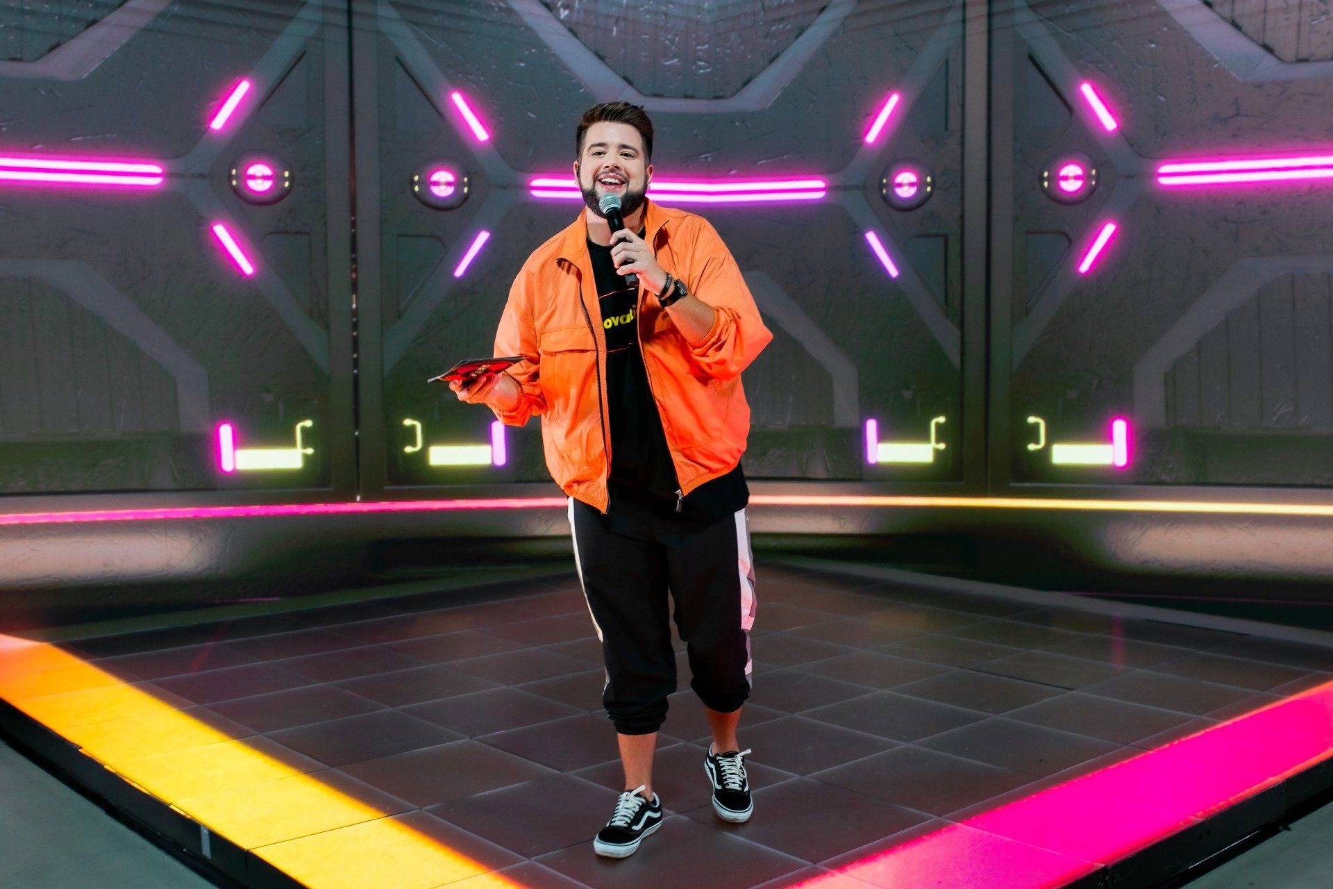 Um homem com uma jaqueta laranja está em pé no palco segurando um microfone.
