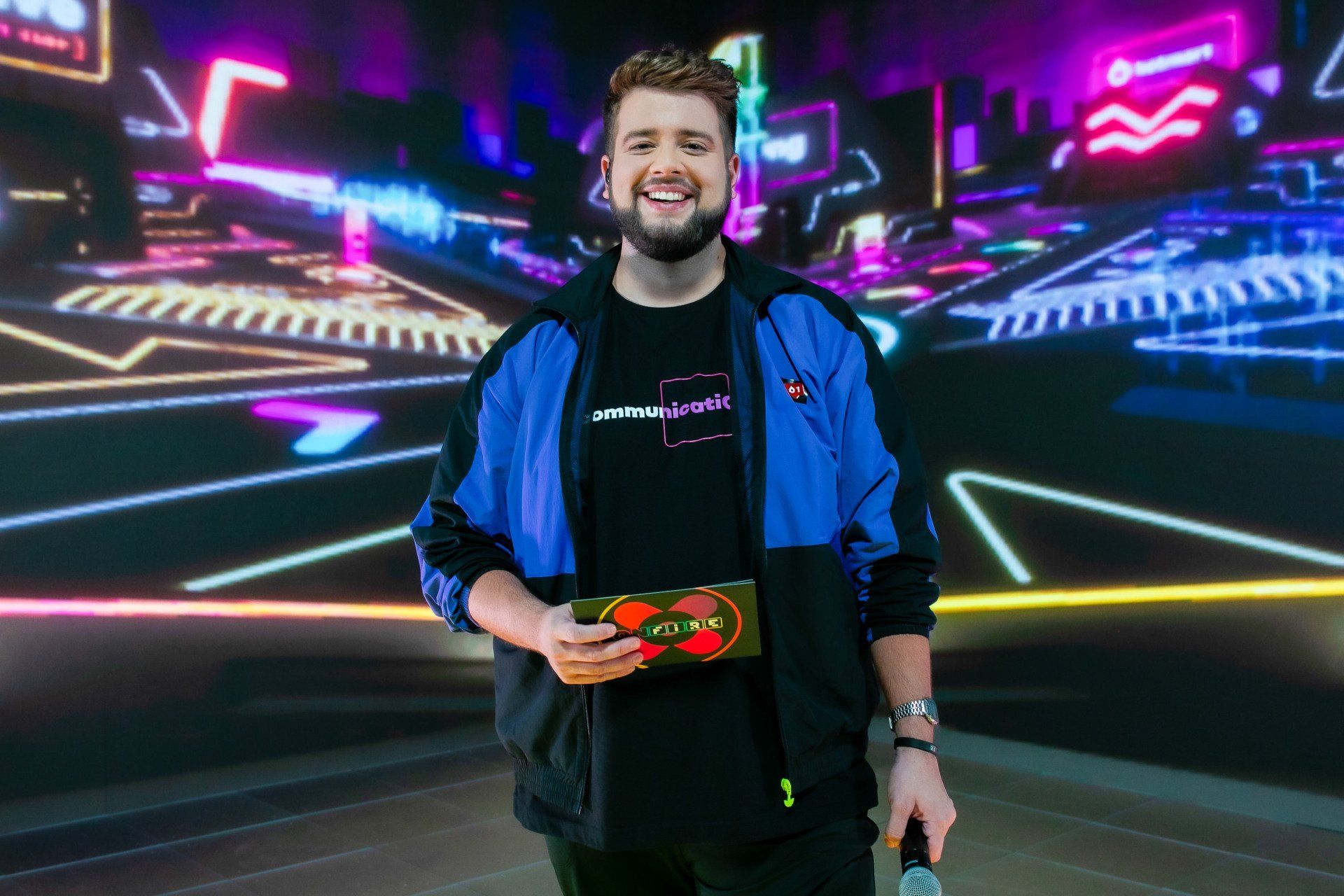 Um homem de jaqueta azul segura um microfone em frente a um fundo neon.