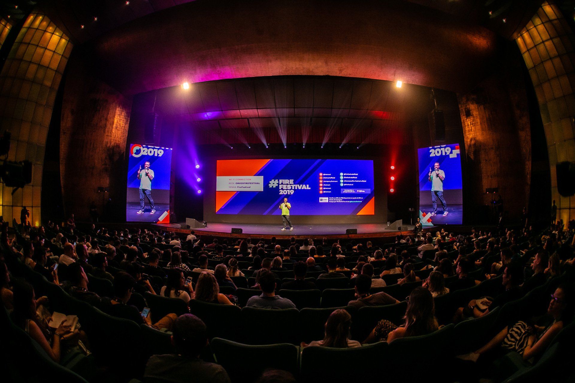 Uma grande multidão de pessoas está sentada em um teatro assistindo a uma apresentação no palco.