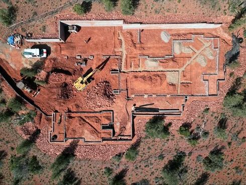 Overhead view of a house foundation under construction on reddish soil; excavator, materials, and equipment visible.