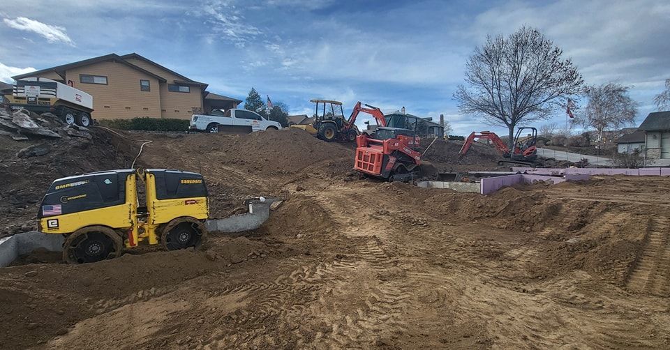 Construction site with heavy machinery like excavators and a compactor, working on a sloped dirt area with houses in the background.