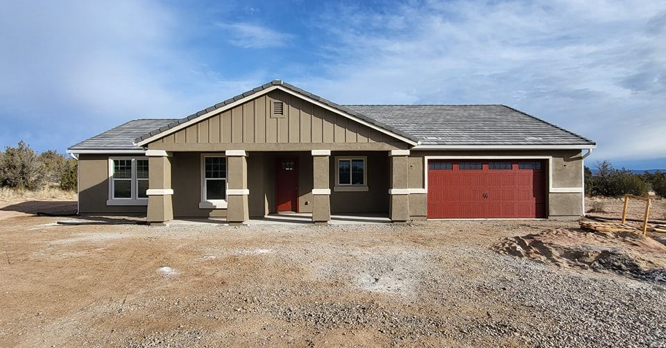 Tan stucco house with red garage door, sitting in a dirt lot under a blue sky.