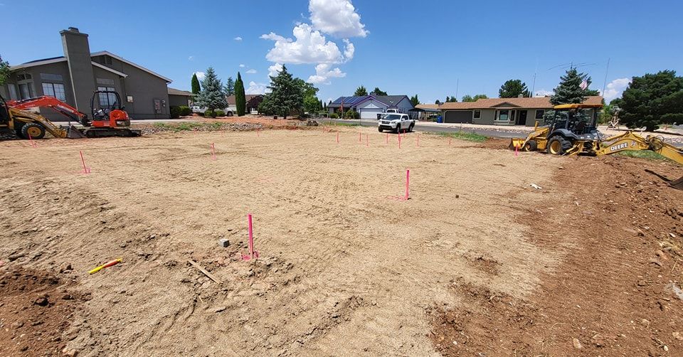 Construction site with leveled dirt, pink stakes, and heavy machinery, in a residential neighborhood on a sunny day.