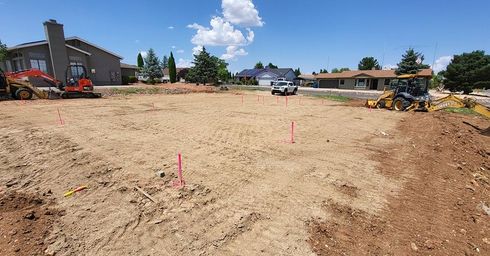 Construction site with leveled dirt, pink stakes, and heavy machinery, in a residential neighborhood on a sunny day.
