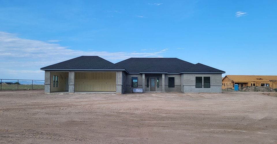 A house under construction with a dark roof and unfinished exterior, set in a dry, open area.