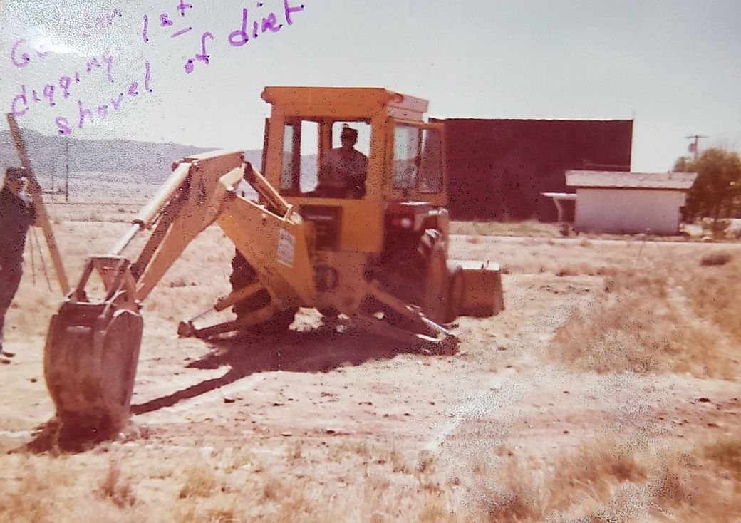 Yellow backhoe digging dirt outdoors. Person inside cab. Brown building, small white building, and mountains in background.