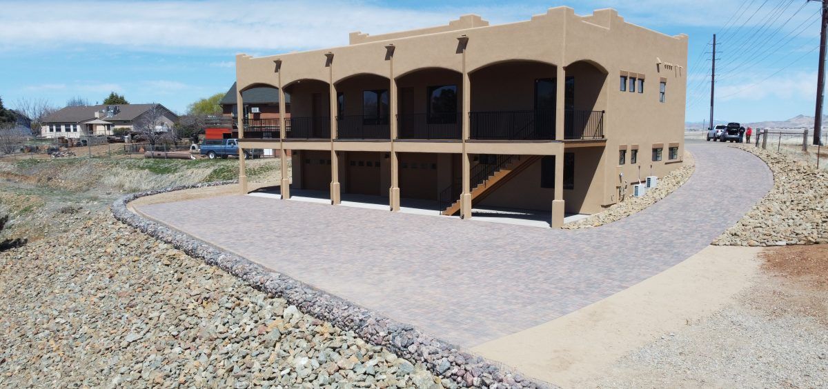 Two-story tan building with a gravel driveway and retaining wall on a hillside.