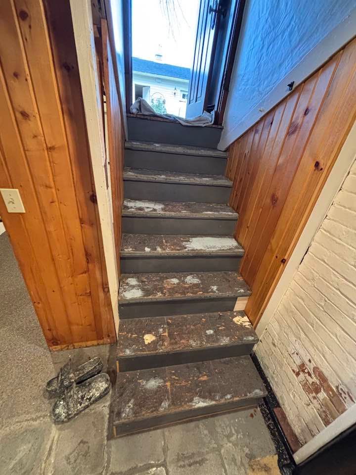 Staircase with worn, dark steps, surrounded by wood paneling and peeling paint. Gray stone flooring.