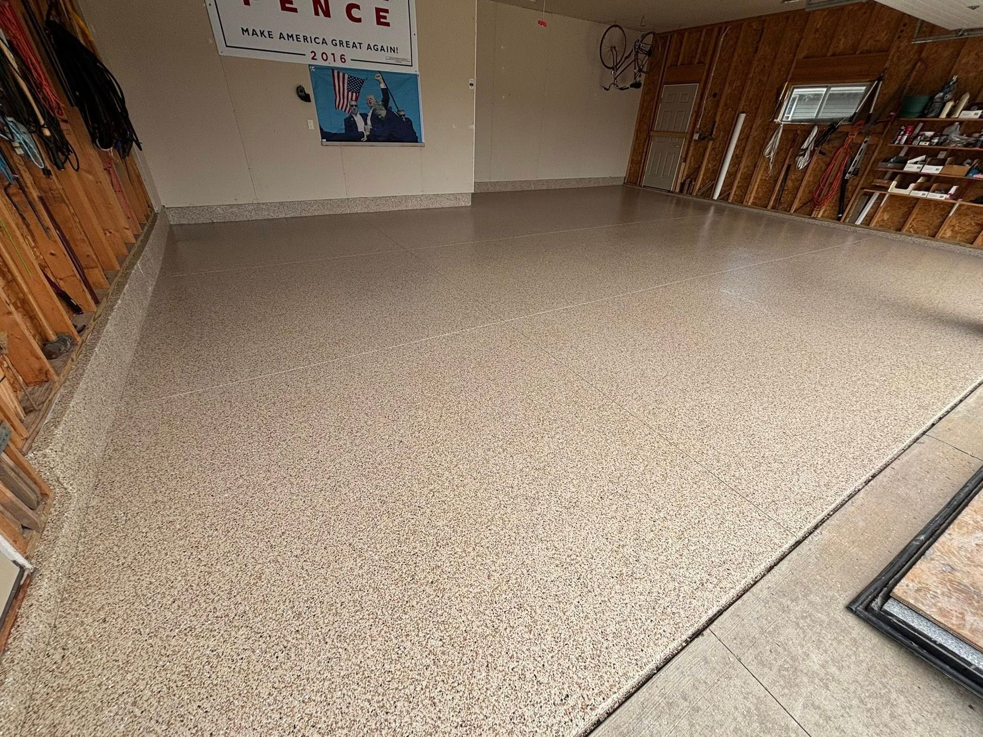 Garage with a speckled epoxy floor. Walls are tan, framing is visible. A poster hangs on the wall.