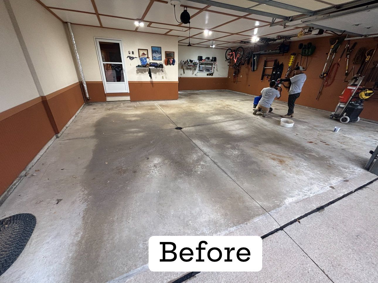 Garage interior, before renovation. Concrete floor, two people, various tools and items on walls.