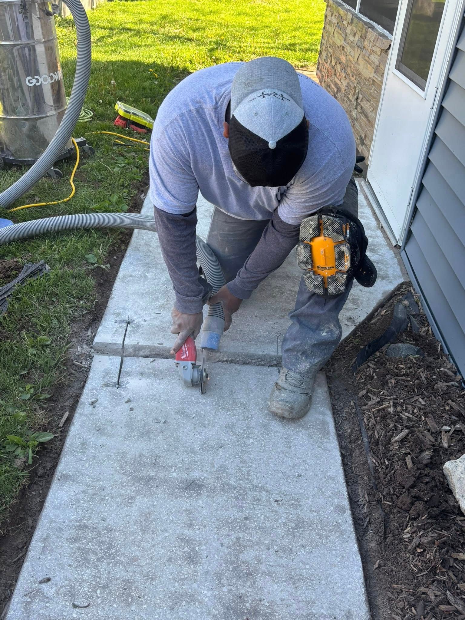 Man kneeling, using a tool on a concrete sidewalk. Gray shirt, gray hat, near building, green grass.
