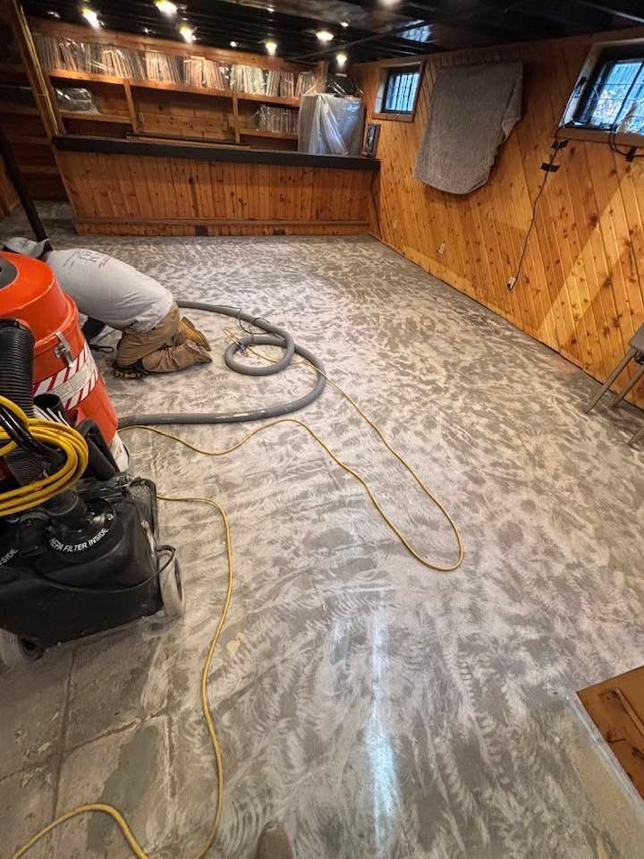 Person grinding a concrete floor in a basement. The floor is light grey and the walls are wood paneling.