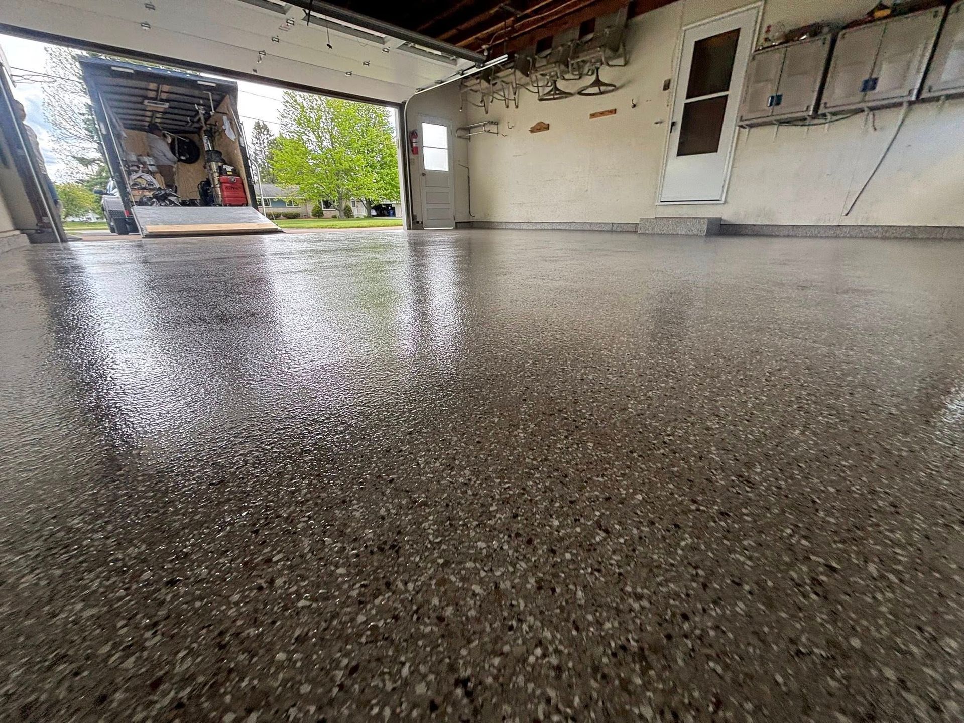 A garage with a shiny, speckled epoxy floor. The garage door is open, revealing a view of the outside.
