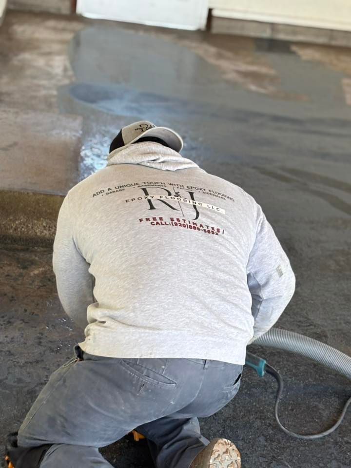 Person kneeling, using a grinder on a concrete floor, wearing a gray hoodie and baseball cap.