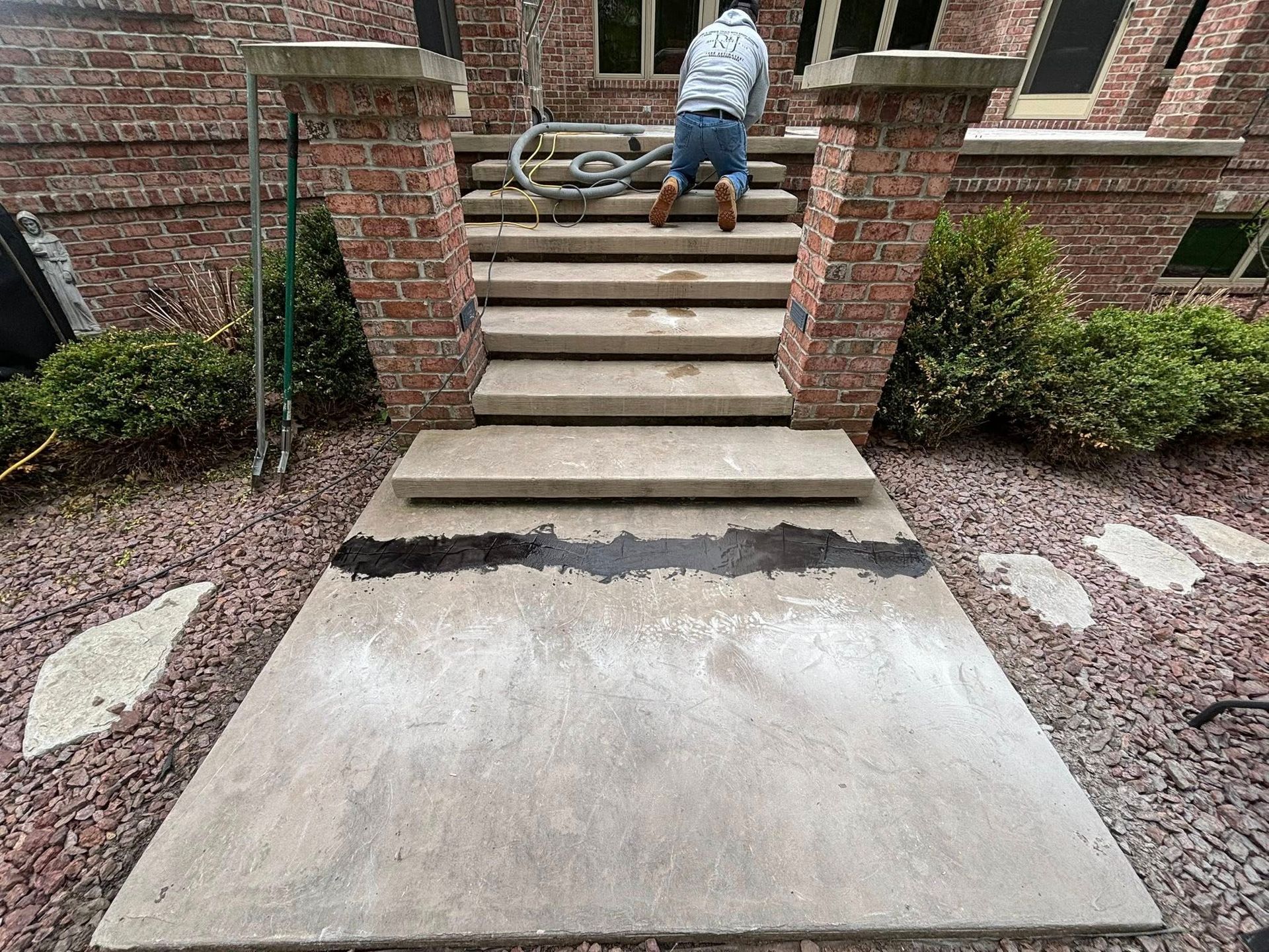 Person working on concrete steps. Brick home with reddish-brown mulch. Partially cleaned, dark section on sidewalk.