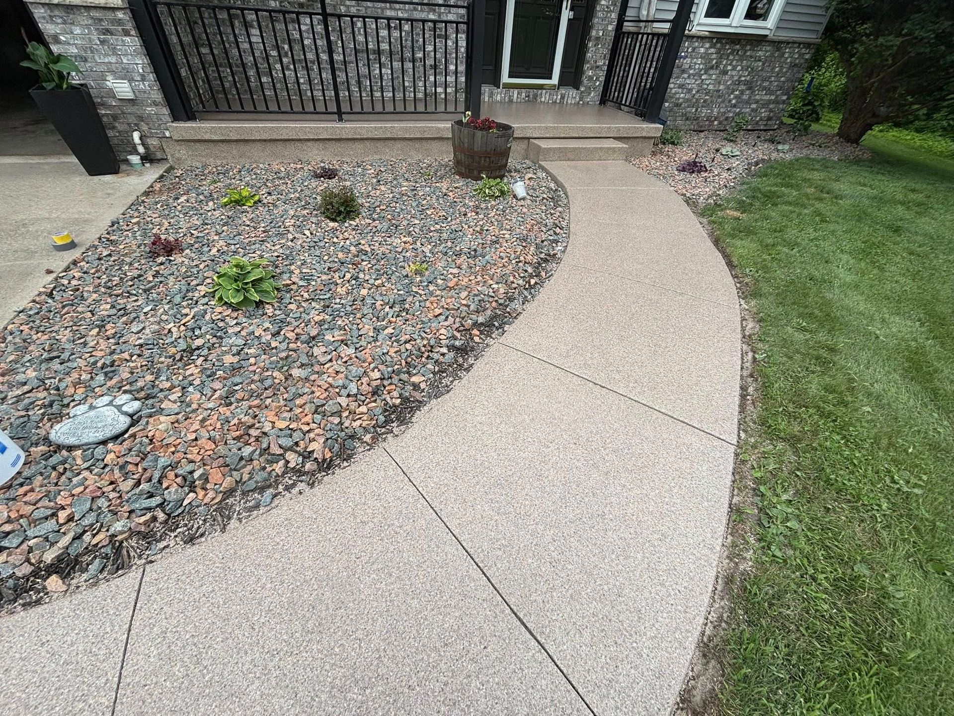 Curving concrete walkway leading to a house entrance, bordered by a rock garden and grass.