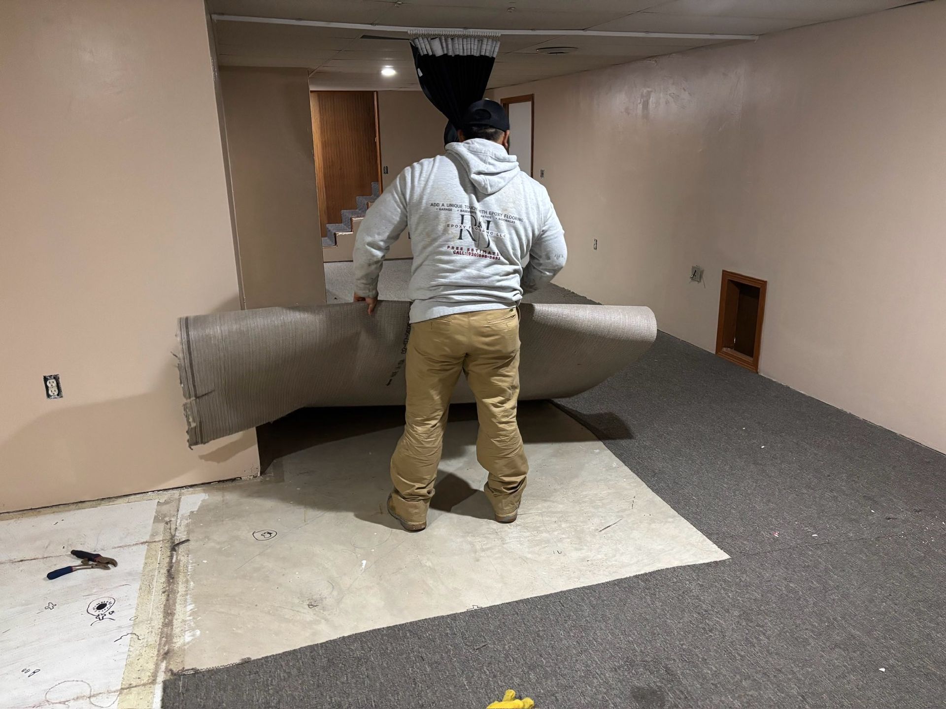 Person rolls out carpet in a basement with tan walls, brown pants, and a gray hoodie.