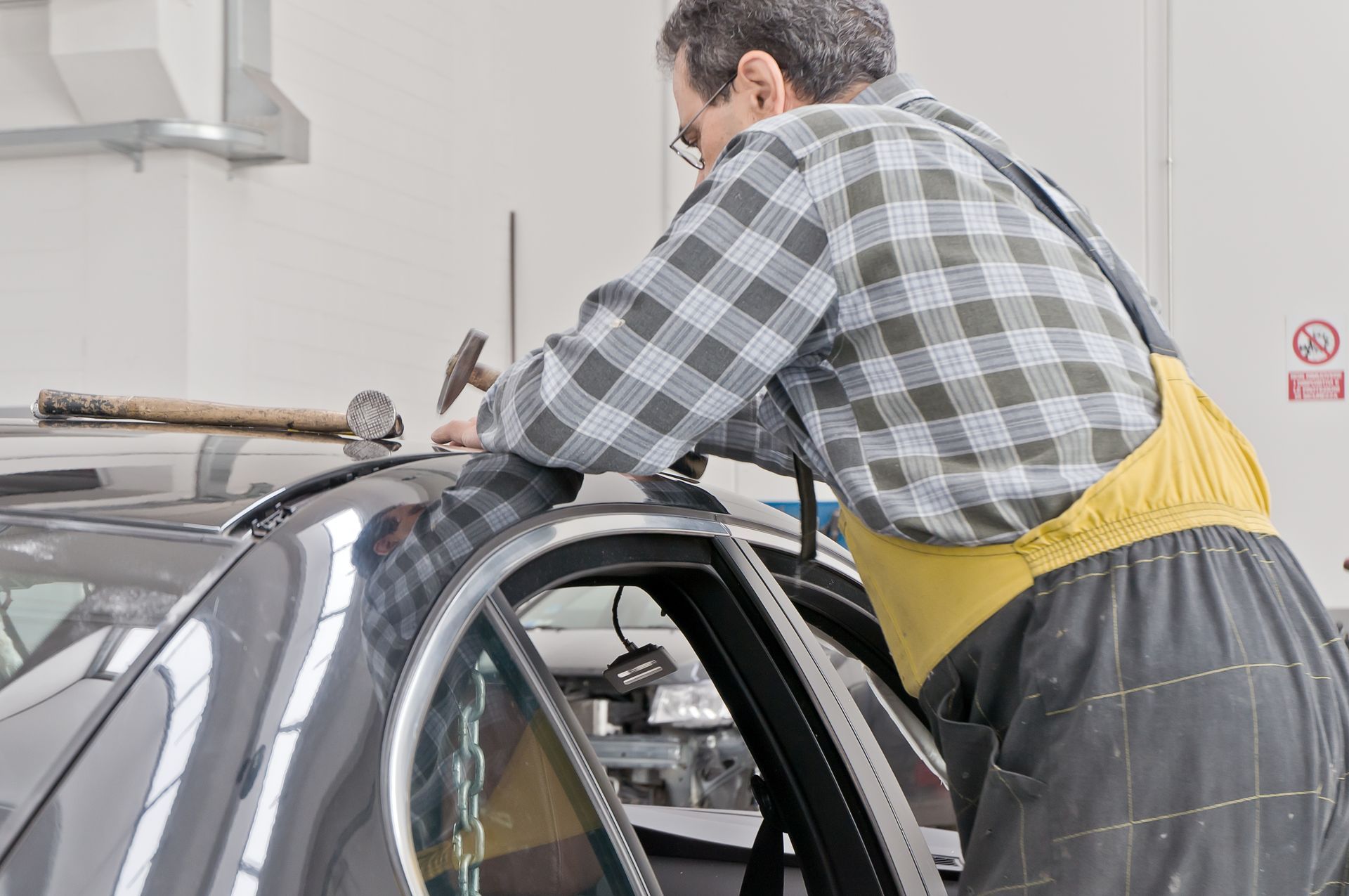 A man in overalls is engaged in auto body hail repair, working diligently on a car in a garage.