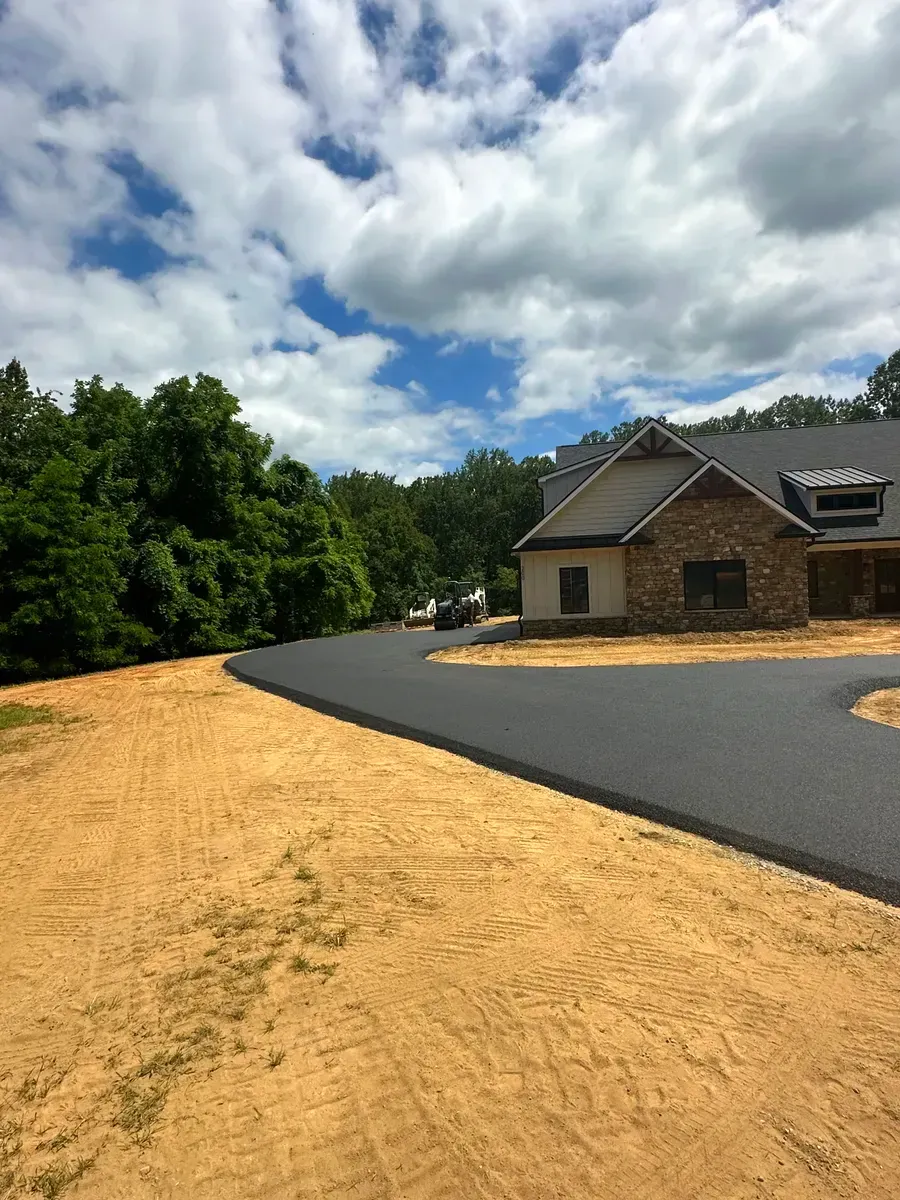 New asphalt driveway curves towards a house with brick and stone facade; sunny day, cloudy sky.