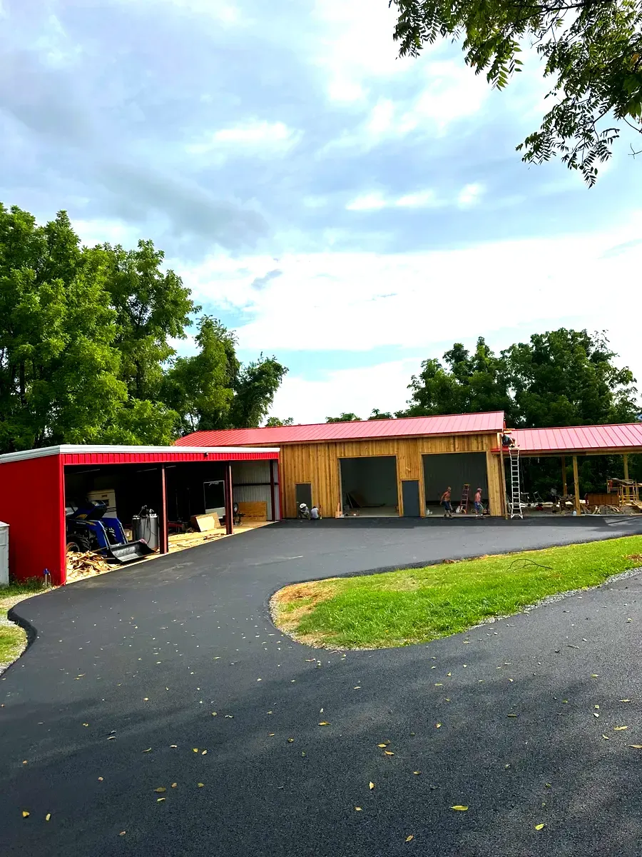 Asphalt driveway leads to a red and wood-colored building with a red roof, surrounded by green trees under a cloudy sky.