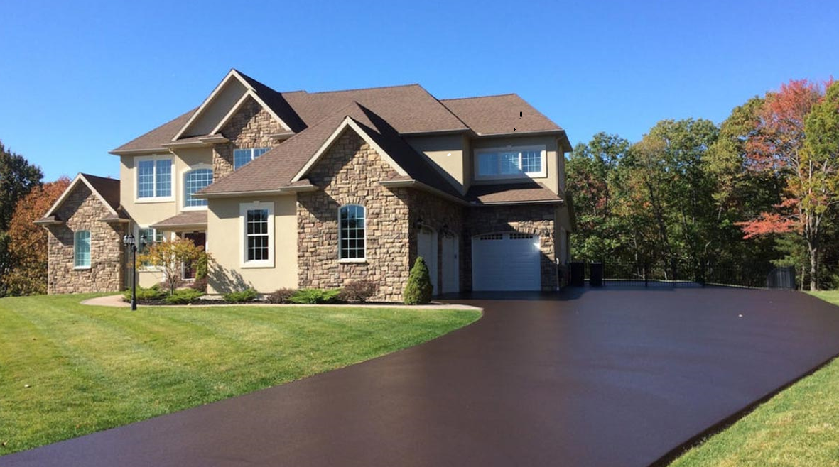 Two-story house with stone facade, tan siding, and a long, black driveway on a sunny day.