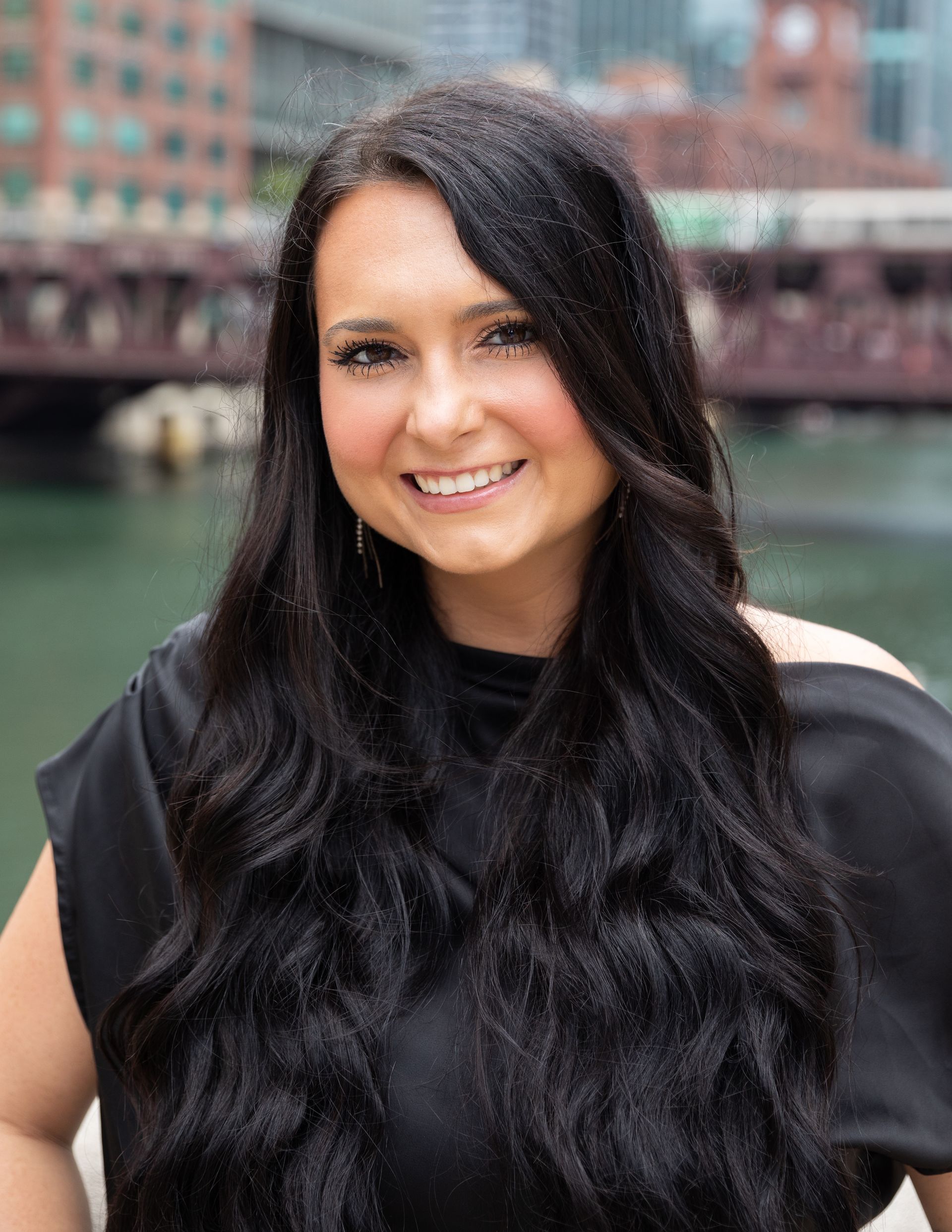 A woman with long black hair is smiling in front of a bridge.