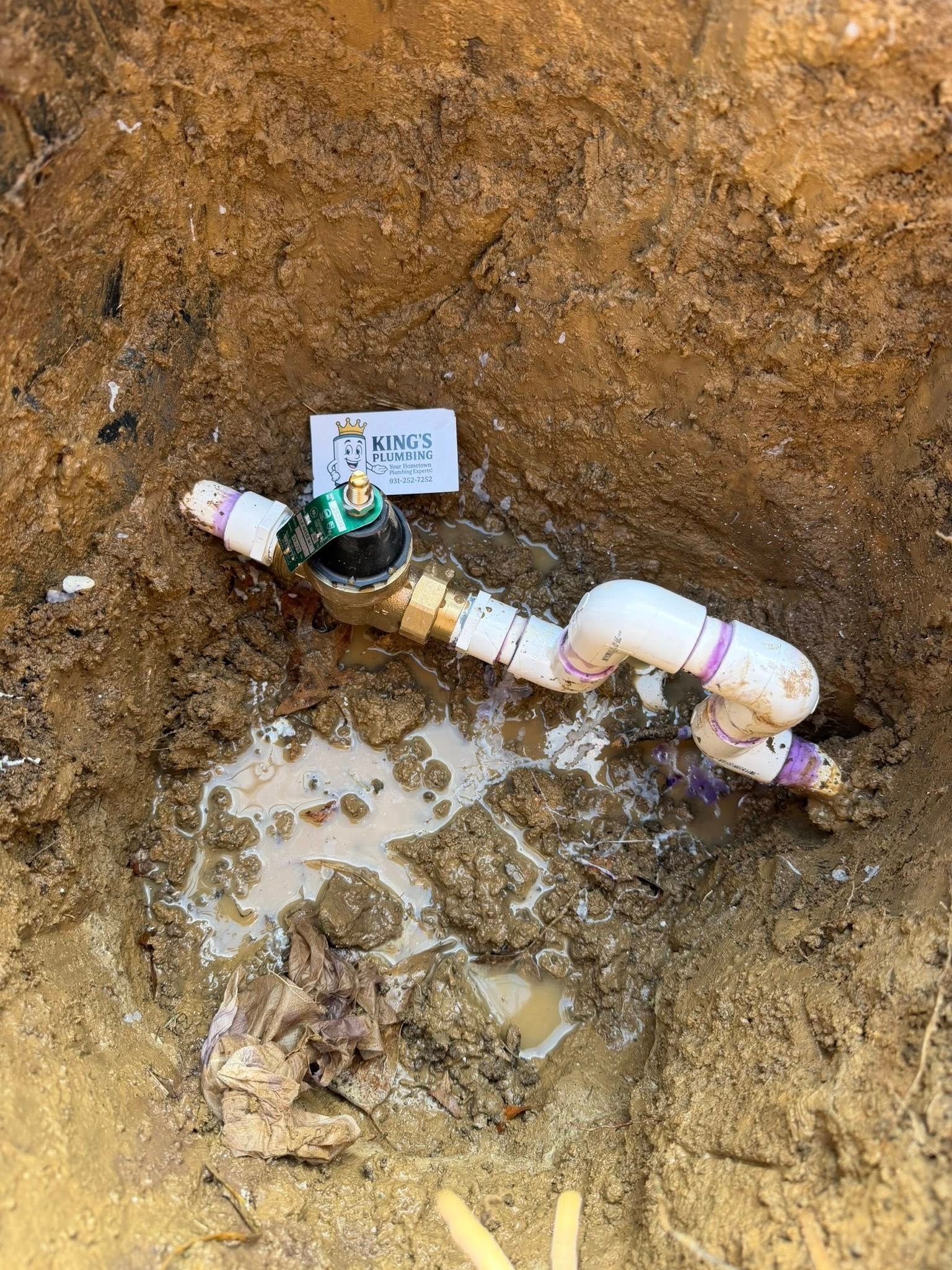 A brass irrigation valve connected to white PVC pipes installed in a deep, muddy dirt trench.