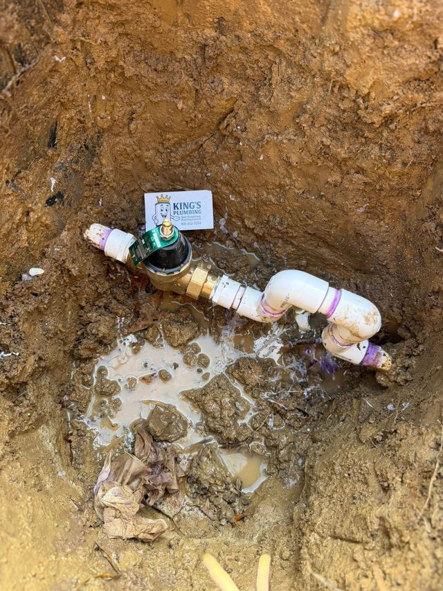 A brass irrigation valve connected to white PVC pipes installed in a deep, muddy dirt trench.