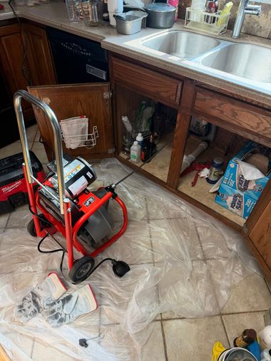 A red motorized drain auger sitting on a plastic sheet in front of an open kitchen cabinet under a sink.