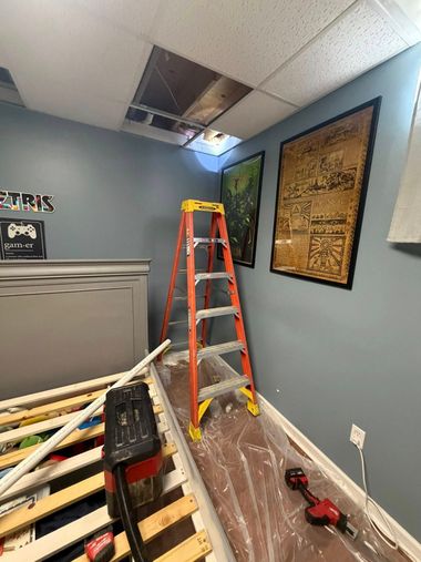 A stepladder sits in a blue-walled room under a missing ceiling tile, with construction tools on the floor nearby.