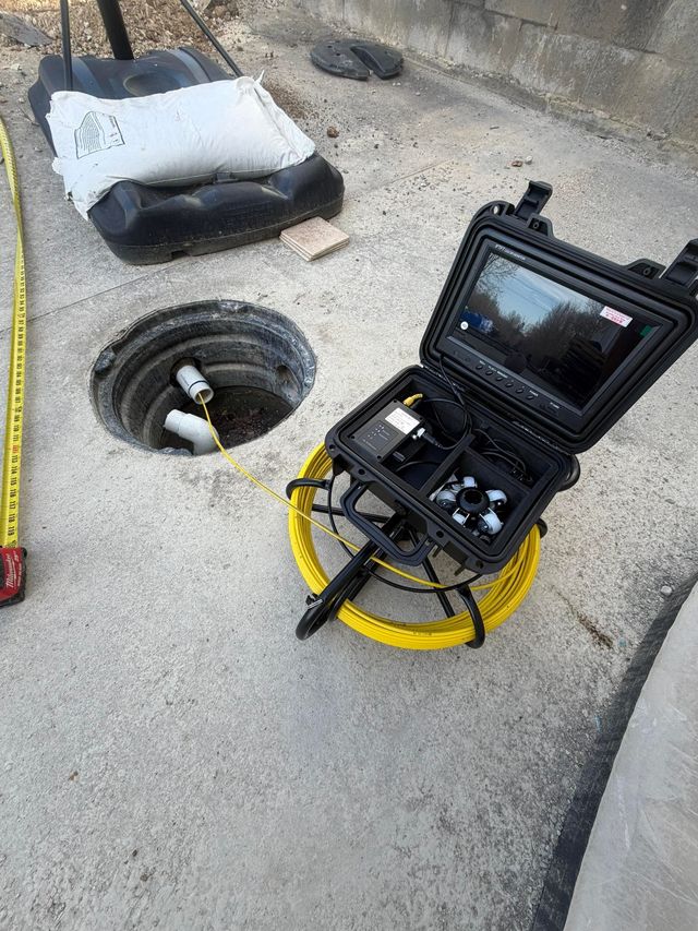 A sewer inspection camera on a yellow reel is positioned next to an open access pipe in a concrete ground surface.