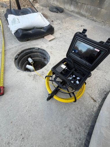 A sewer camera system with a yellow cable reel and a monitor, set up on concrete near an open pipe access point.