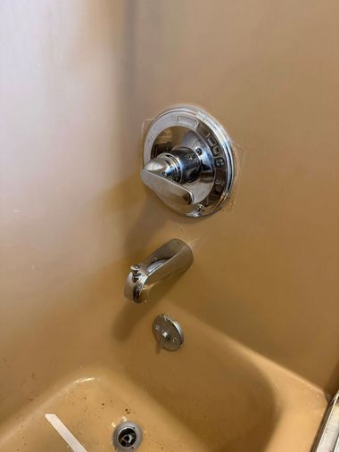 A chrome shower handle, spout with a diverter, and drain set against a tan-colored bathtub wall.