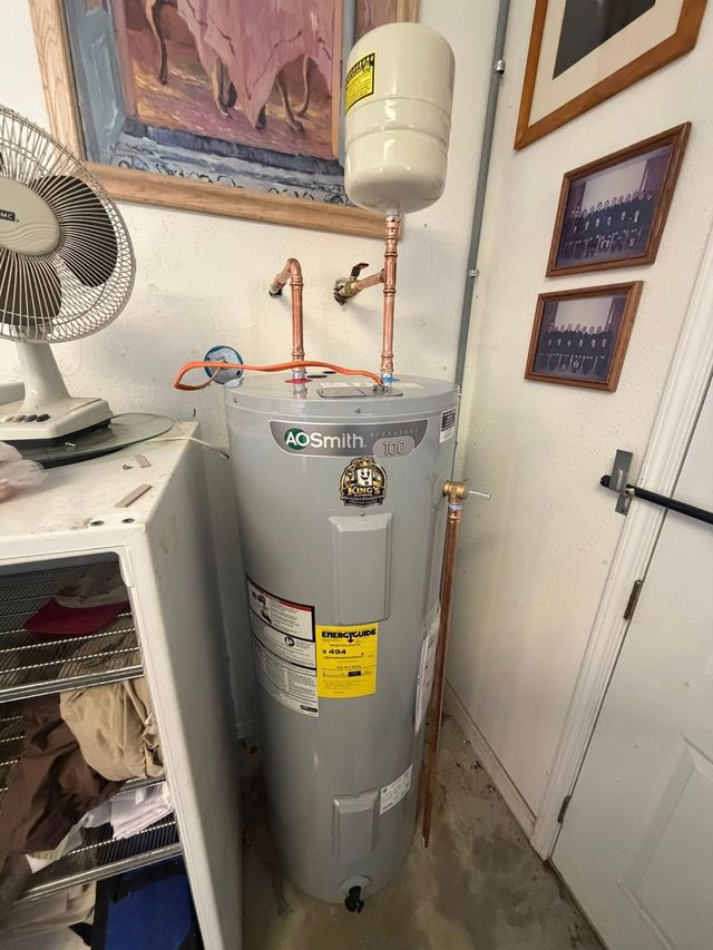 A grey A.O. Smith water heater with an attached expansion tank, standing in a room with a fan, shelf, and framed art.
