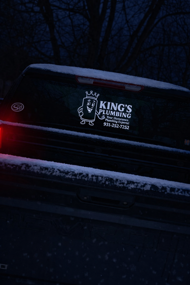 A white decal on a snowy pickup truck rear window for King's Plumbing, featuring a cartoon character wearing a crown.