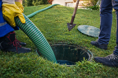 Two people work on a septic tank in a grassy yard, using a large green suction hose and a tool to move the cover.