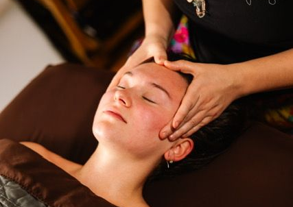 Woman receiving head massage, eyes closed, with a man in the background also receiving a massage, in a spa setting.